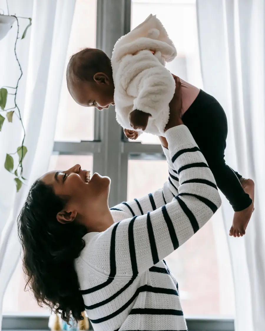 A mother lifts her baby overhead by a window
